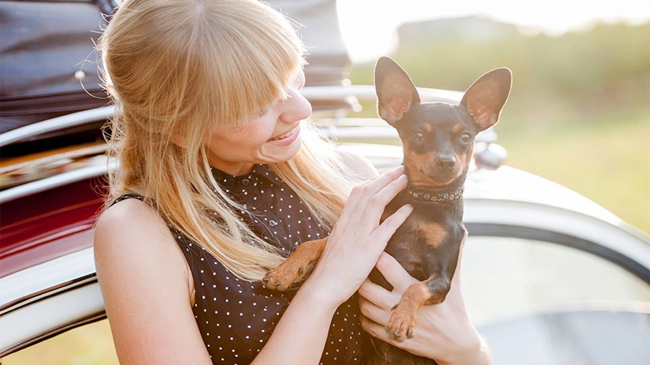A young woman holding her small dog standing in front of her car