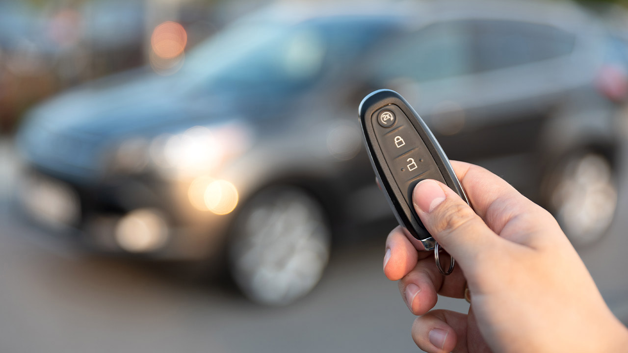A close up of a hand holding a car key fob with the thumb depressing the lock button. A car is in the background