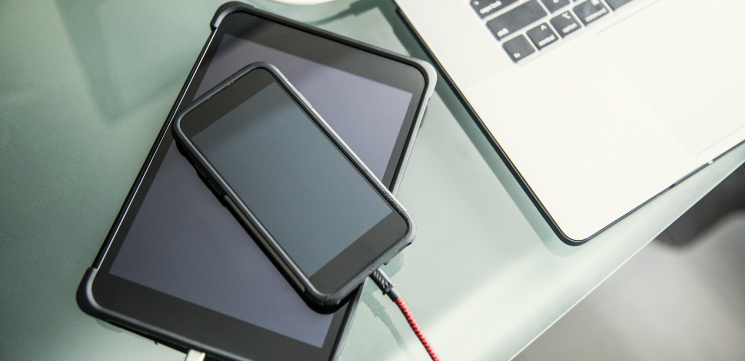 A computer tablet, smartphone and laptop sit on a desk while charging.