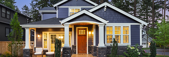Dark blue two-story house with brown front door and porch lights on at dusk.